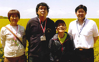 菜の花畑を背景に学園の職員と坂下さん