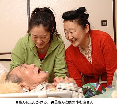 写真:ベッドに寝ている晋さんに、鶴英さんと娘のむぎさんが笑顔で語りかける