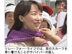 写真:紫色のスカーフを首に巻いてもらうアグネスさん。リレー フォー ライフでは、紫色のスカーフはサバイバーの証し。