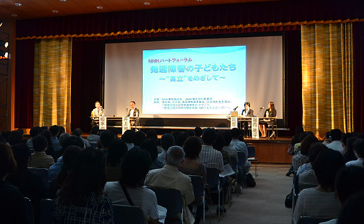 写真：フォーラムのステージ上、左から司会の町永さん、井上さん、スクリーンを挟んで、阿部さん、堀内さんの順に並んでいる。