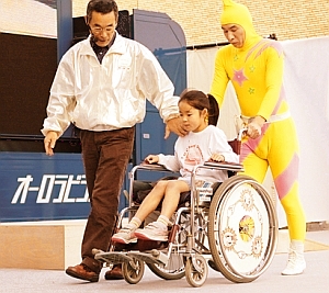 写真:教育テレビのキャラクターが、女の子の乗った車いすを押している
