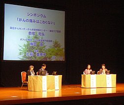 写真:舞台全体。スクリーンの下に4人の出演者が並んで座る。