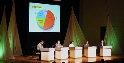 写真:舞台全景。講師が4人並ぶ。中央にスクリーン。