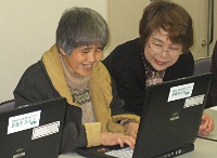写真：パソコンを操作する視覚障害のある人。横には見える人が座って使い方をサポートしている。